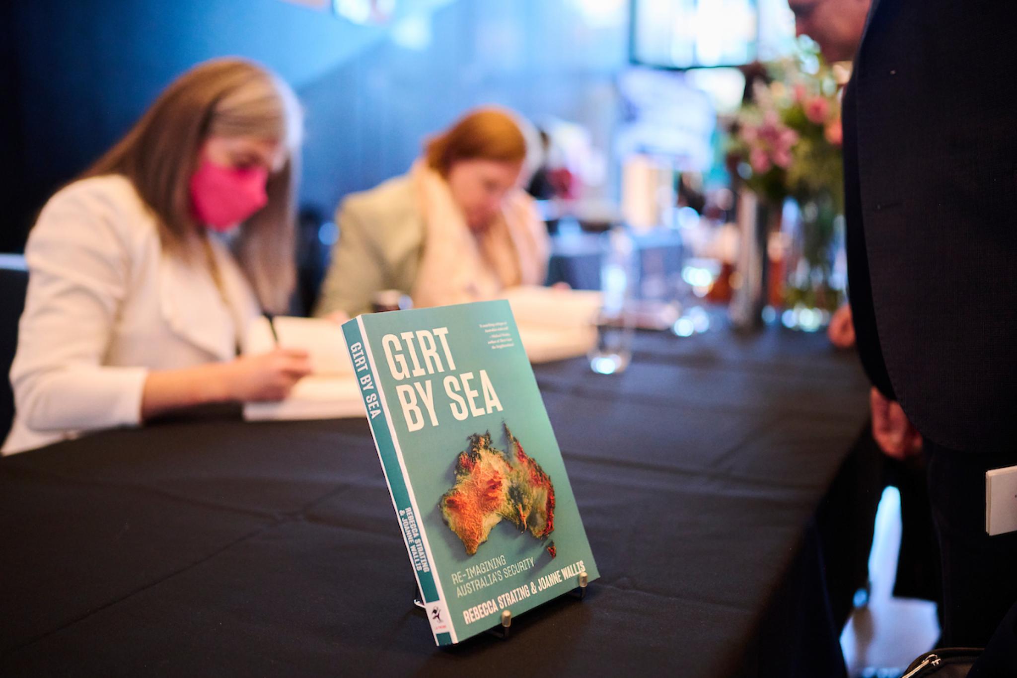 Girt by sea book on table, with authors signing books in background